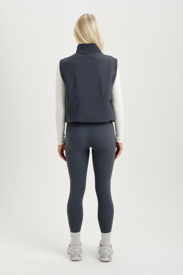 Woman turned around wearing a white long sleeve and gray vest on a white background  