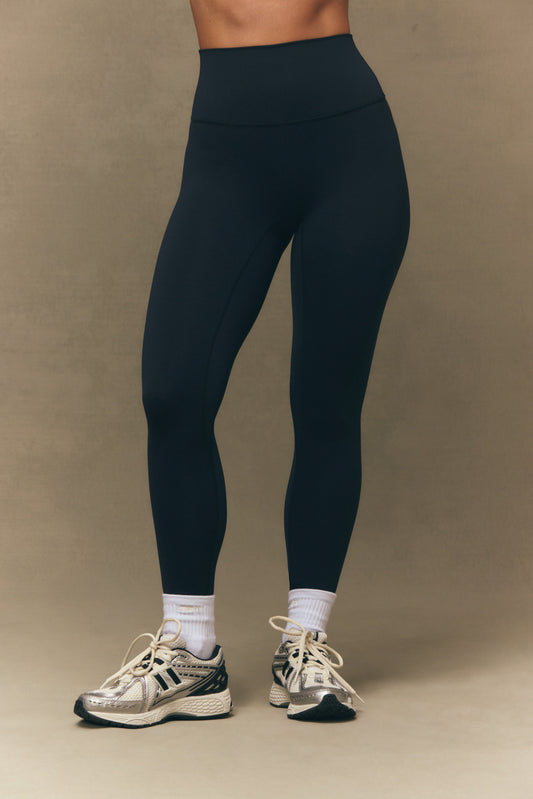 Person wearing black leggings and white sneakers on a beige background <model-info>Bre wears a size S and is 5’7”, 25” waist, 32" bust.</model-info>