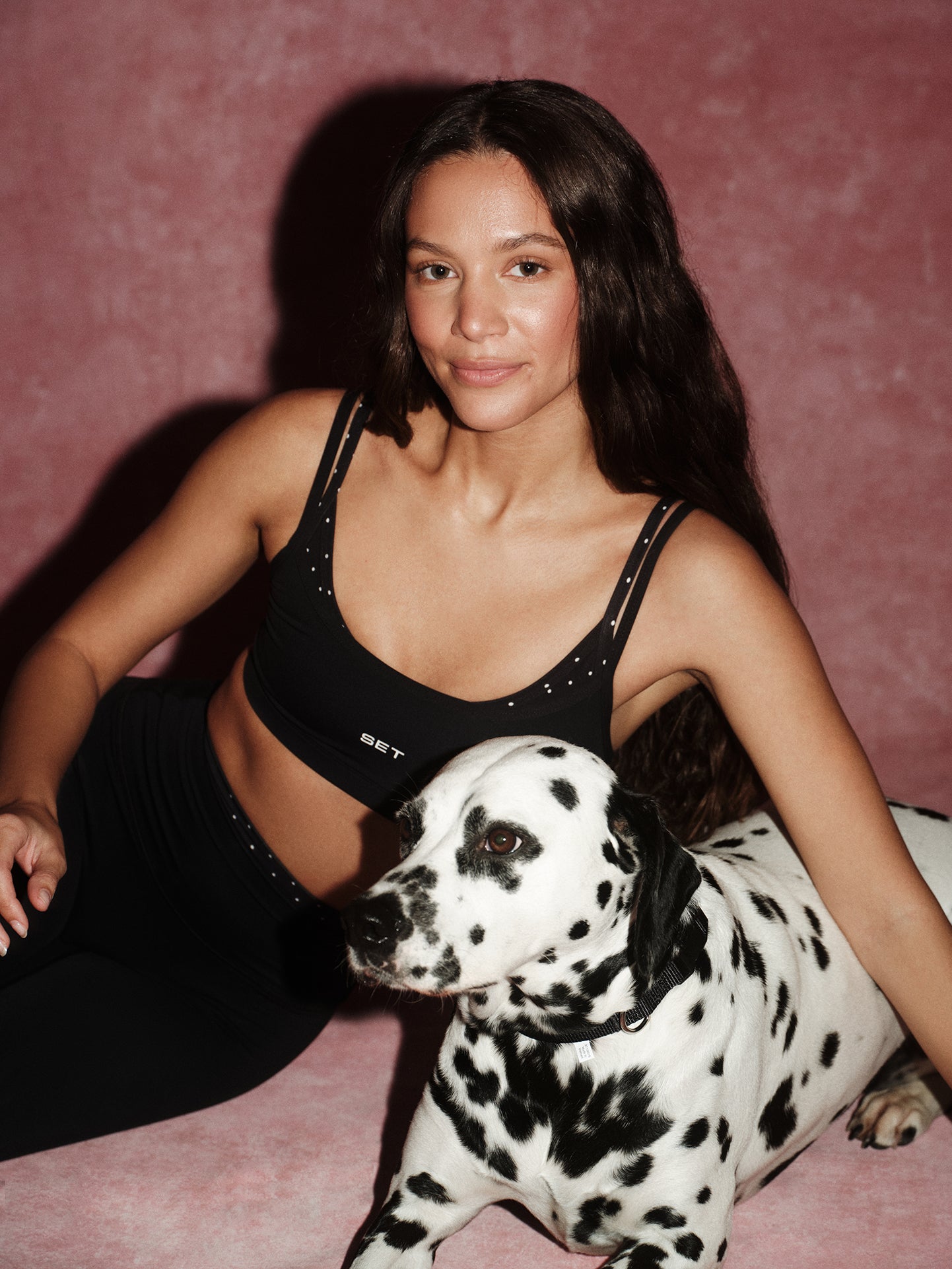 Woman in black sports bra and leggings sitting with a Dalmatian dog on a pink background
