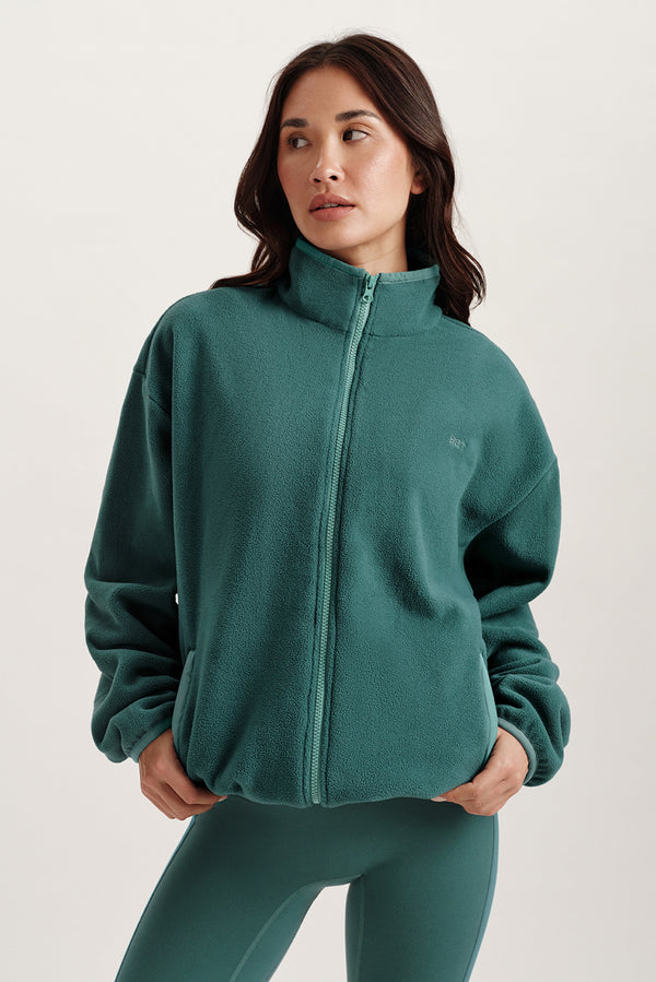 Woman wearing a teal fleece jacket and leggings on a light background 