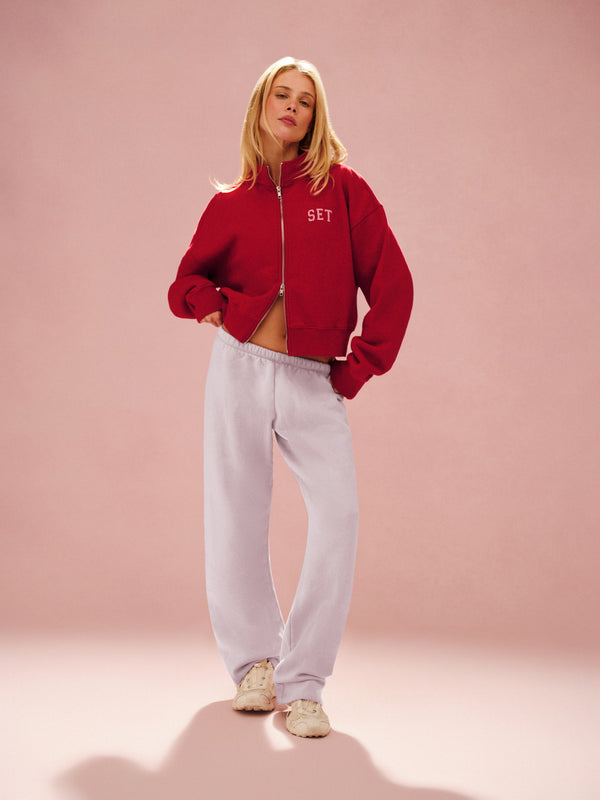 Woman wearing a red jacket with 'SET' logo and light gray pants on a pink background