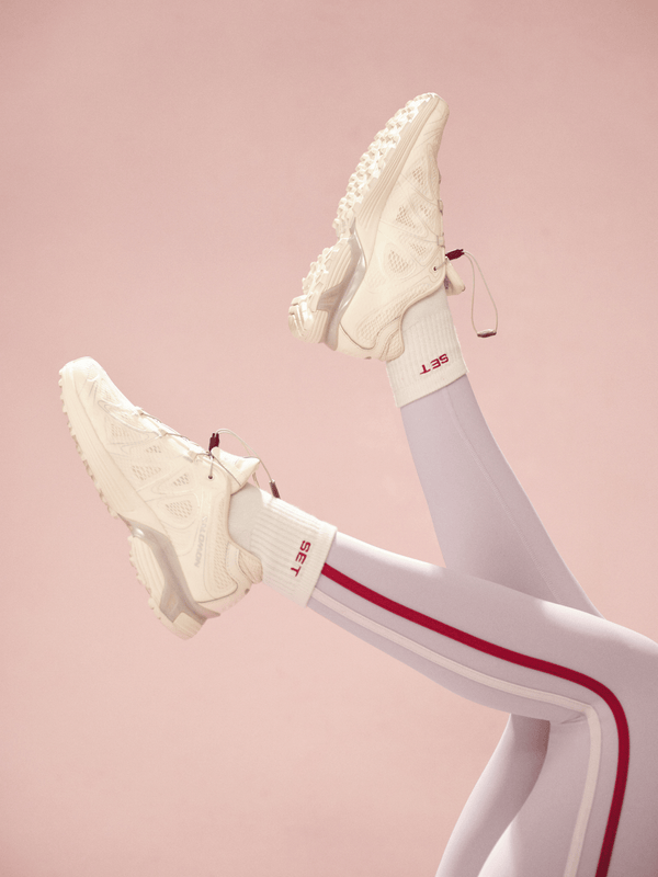 White sneakers with white socks with red accents worn with light purple leggings with stripes on a pink background 