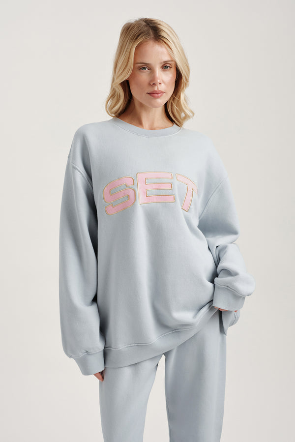 Woman wearing a light blue sweatshirt with 'SET' printed in pink on a plain background