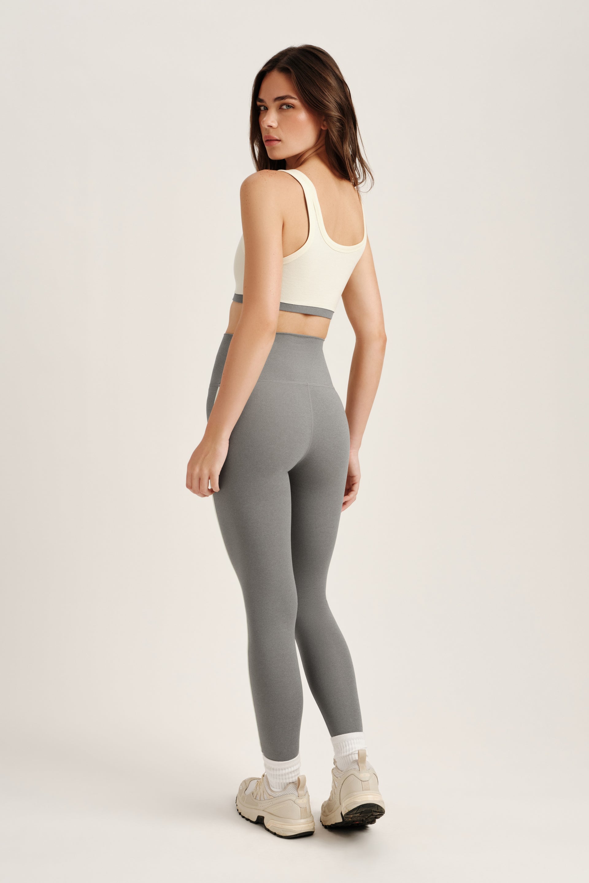 Woman wearing a white sports bra and gray leggings on a light gray background