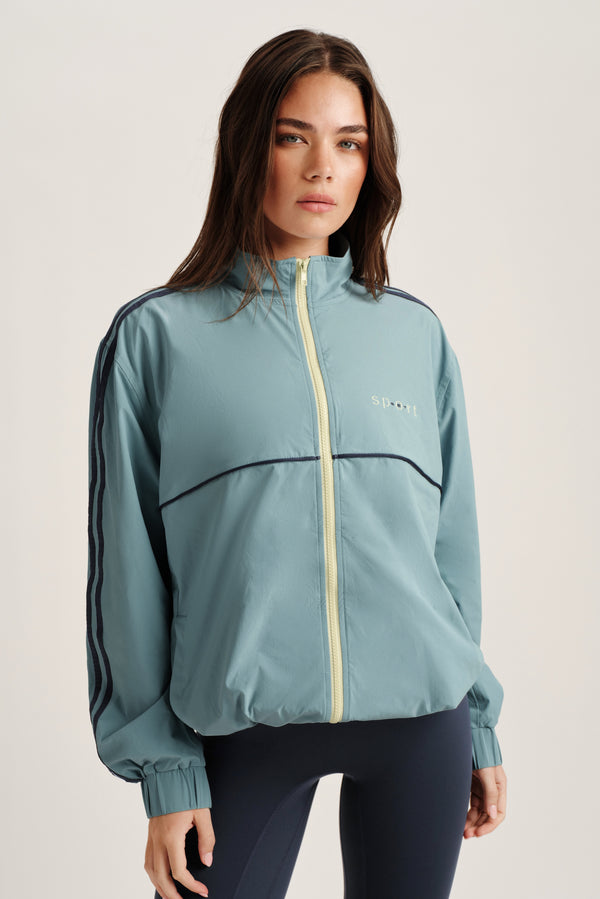 Woman wearing a light blue athletic jacket with a brand logo on a plain background 