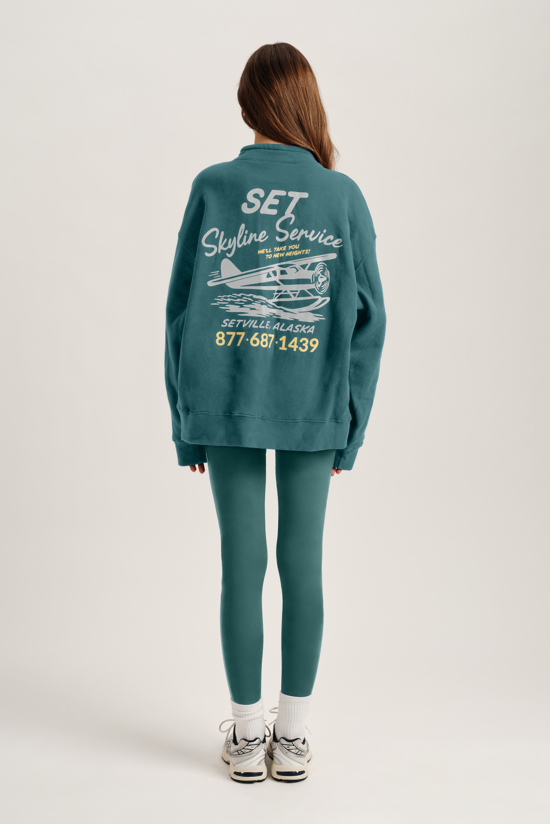 Person wearing a teal sweatshirt with graphics and text on the back, standing against a white background