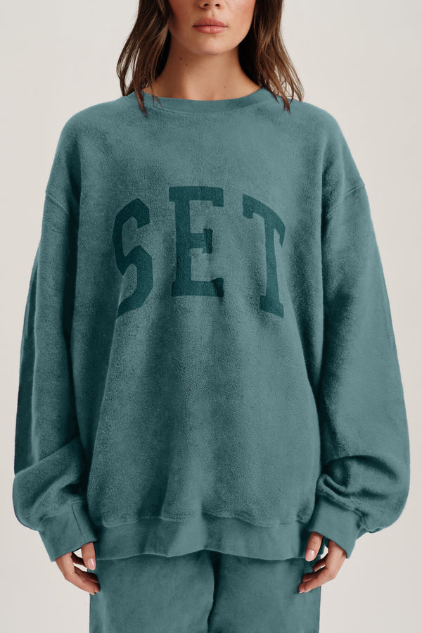 Person wearing a teal sweatshirt with 'SET' printed on it against a neutral background