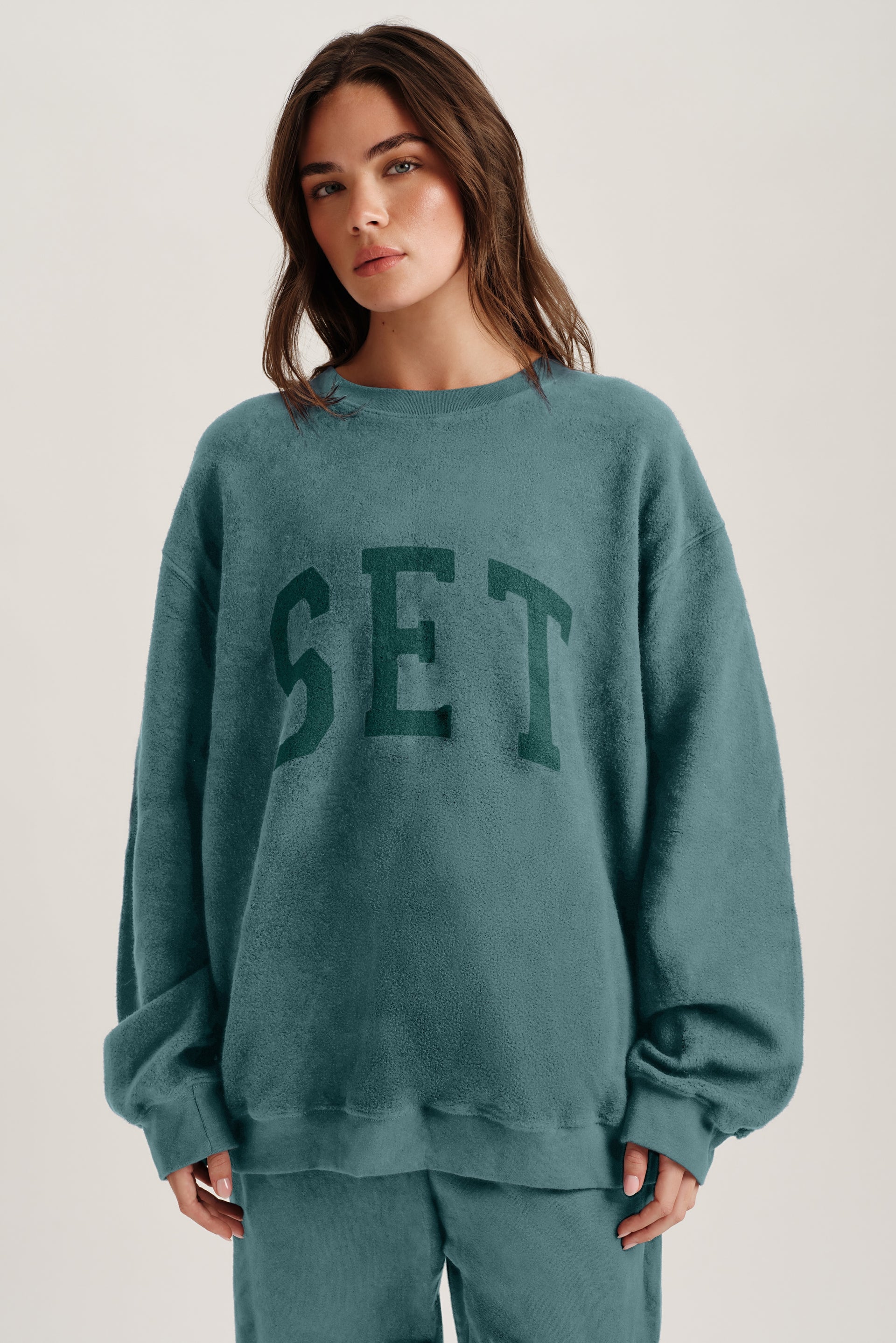 Woman wearing a teal sweatshirt with 'SET' text on a plain background <model-info>Bella wears a size XS/S and is 5’7, 23” waist, 32AB bust.</model-info> 