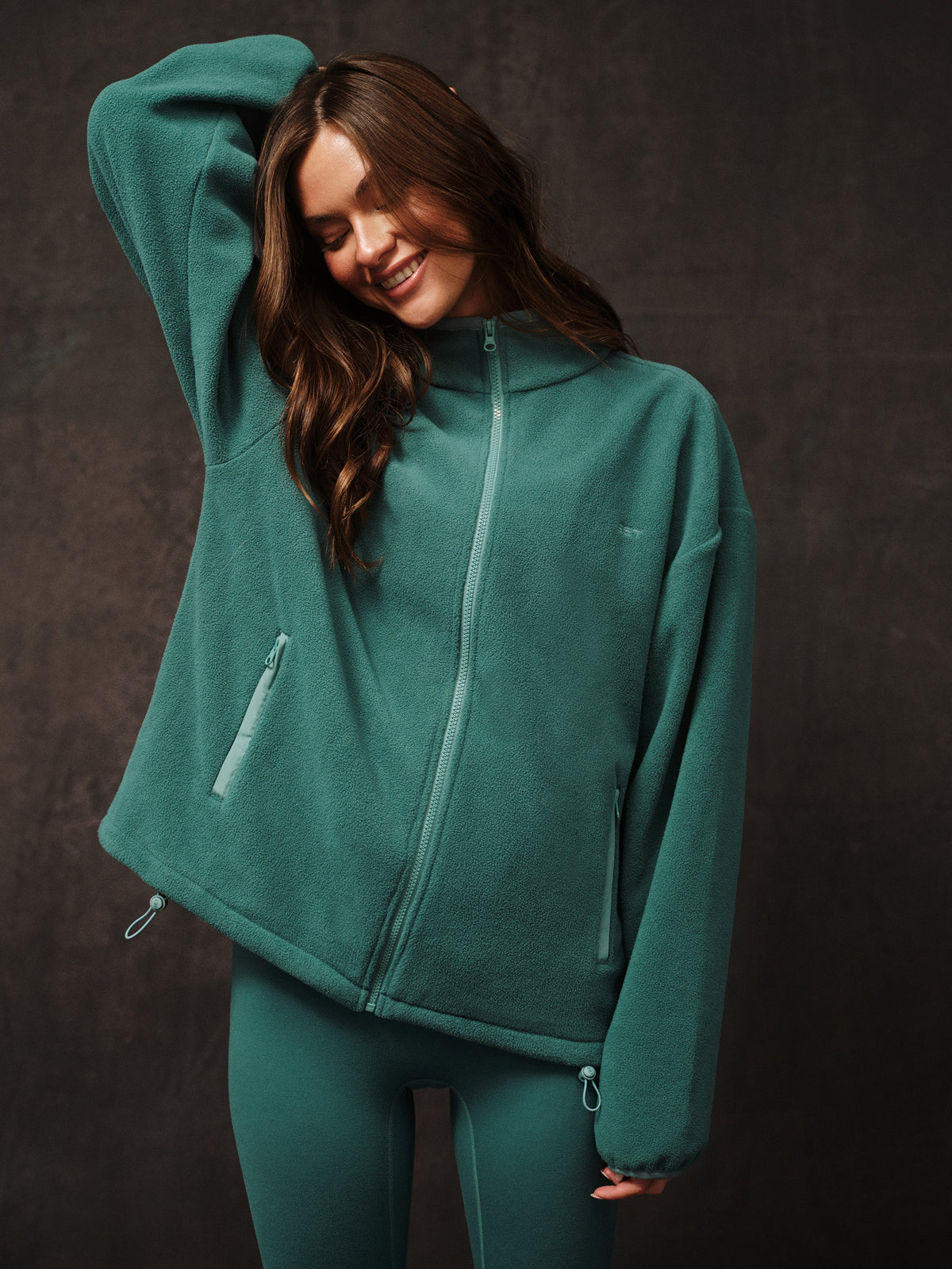 Woman wearing a teal fleece jacket and leggings against a dark background