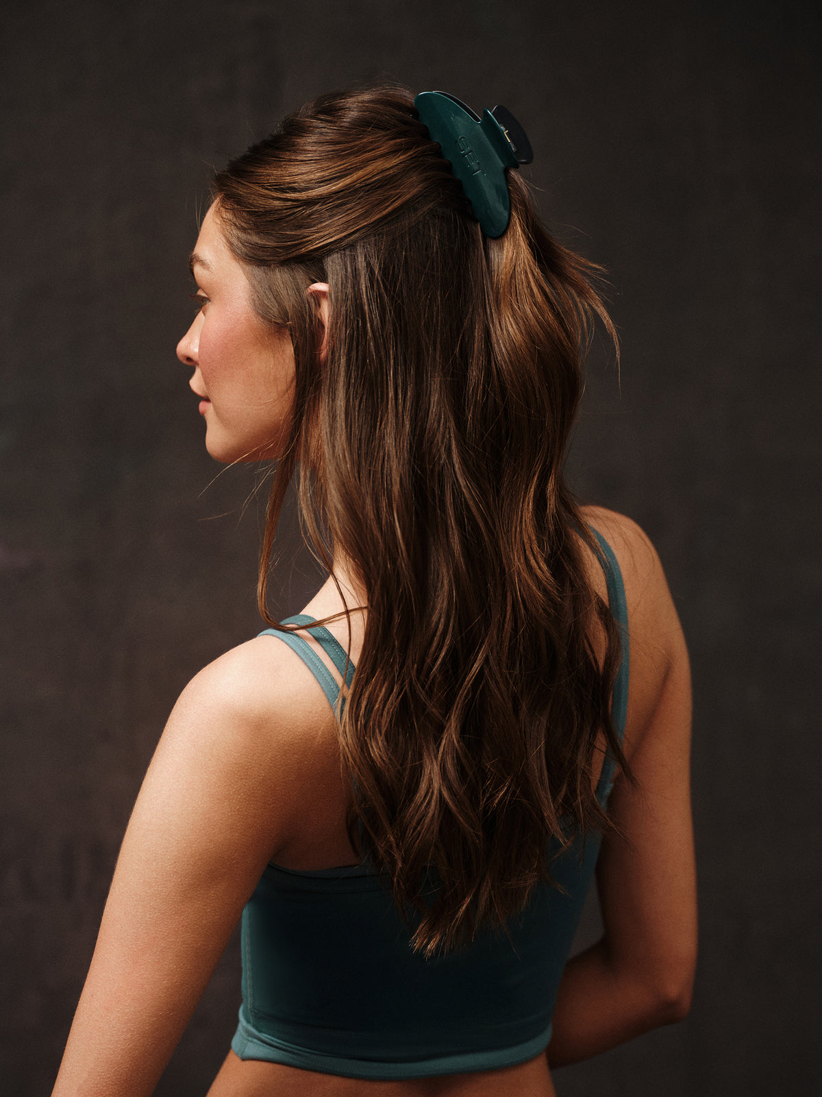 Rear three-quarter view of color block claw clip clipped half-up in model’s hair showing its dark green color and size on a dark studio background