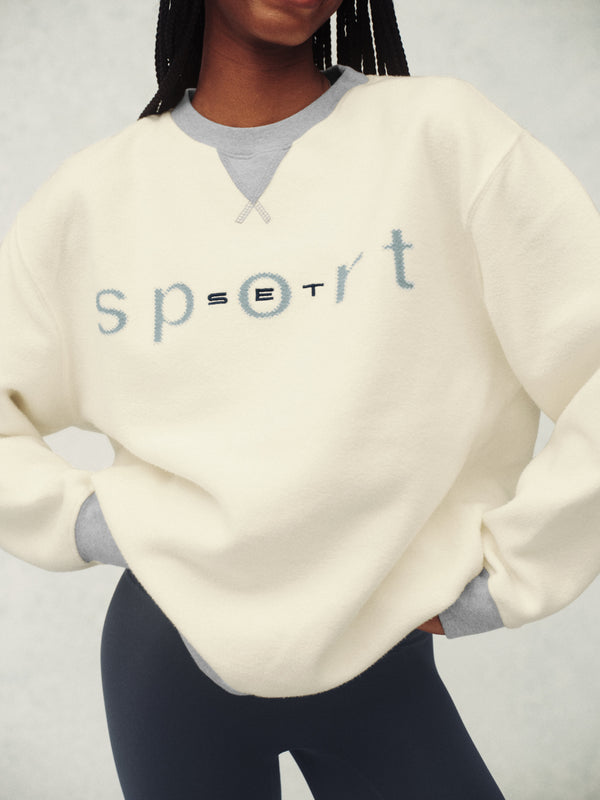 Person wearing a cream-colored sweatshirt with 'sport' text on a plain background
