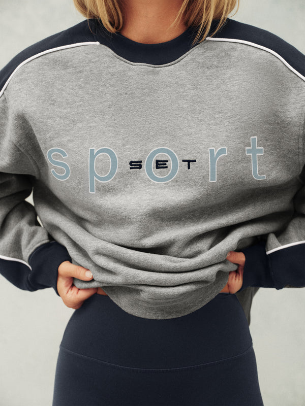 Gray sweatshirt with 'sport' text worn by a person on a neutral background