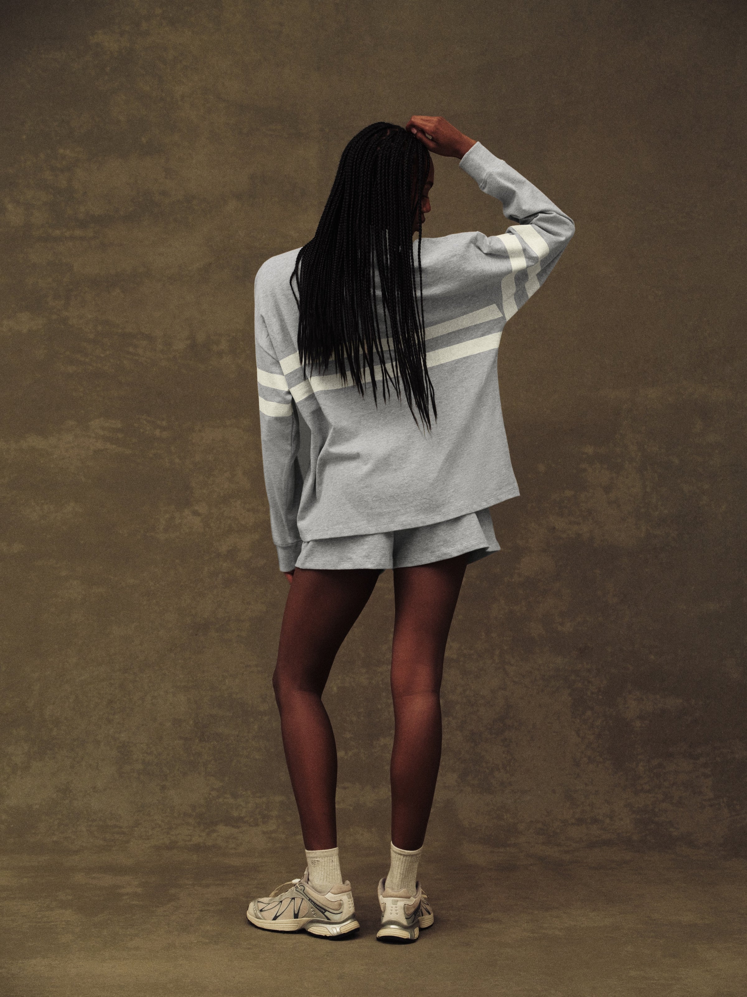 Person wearing a light gray sweater and shorts set with white sneakers against a brown background