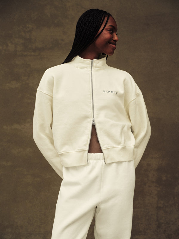 Person wearing a cream-colored tracksuit against a brown background