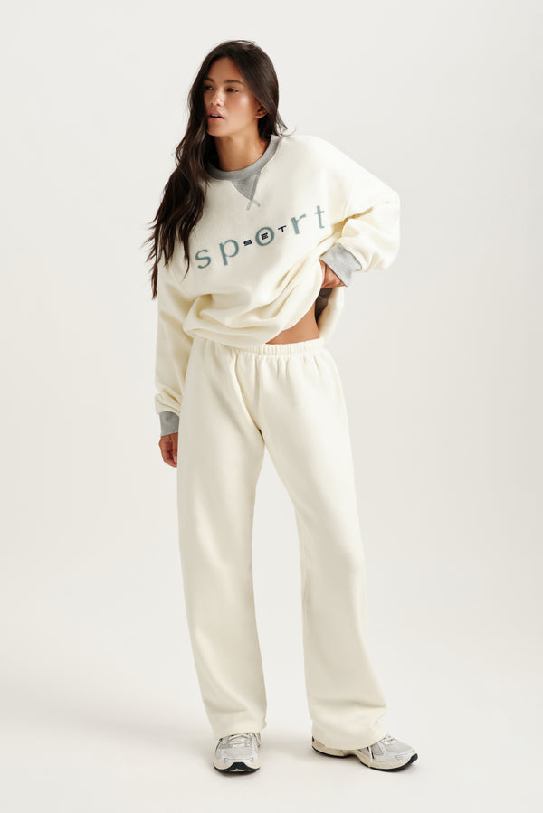 Woman wearing a white sweatshirt with text and cream-colored pants on a white background 