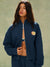 Person wearing a navy blue jacket with a logo on a green background<model-info>Meeya wears a size S and is 5’7, 23” waist, 32B bust.</model-info> 