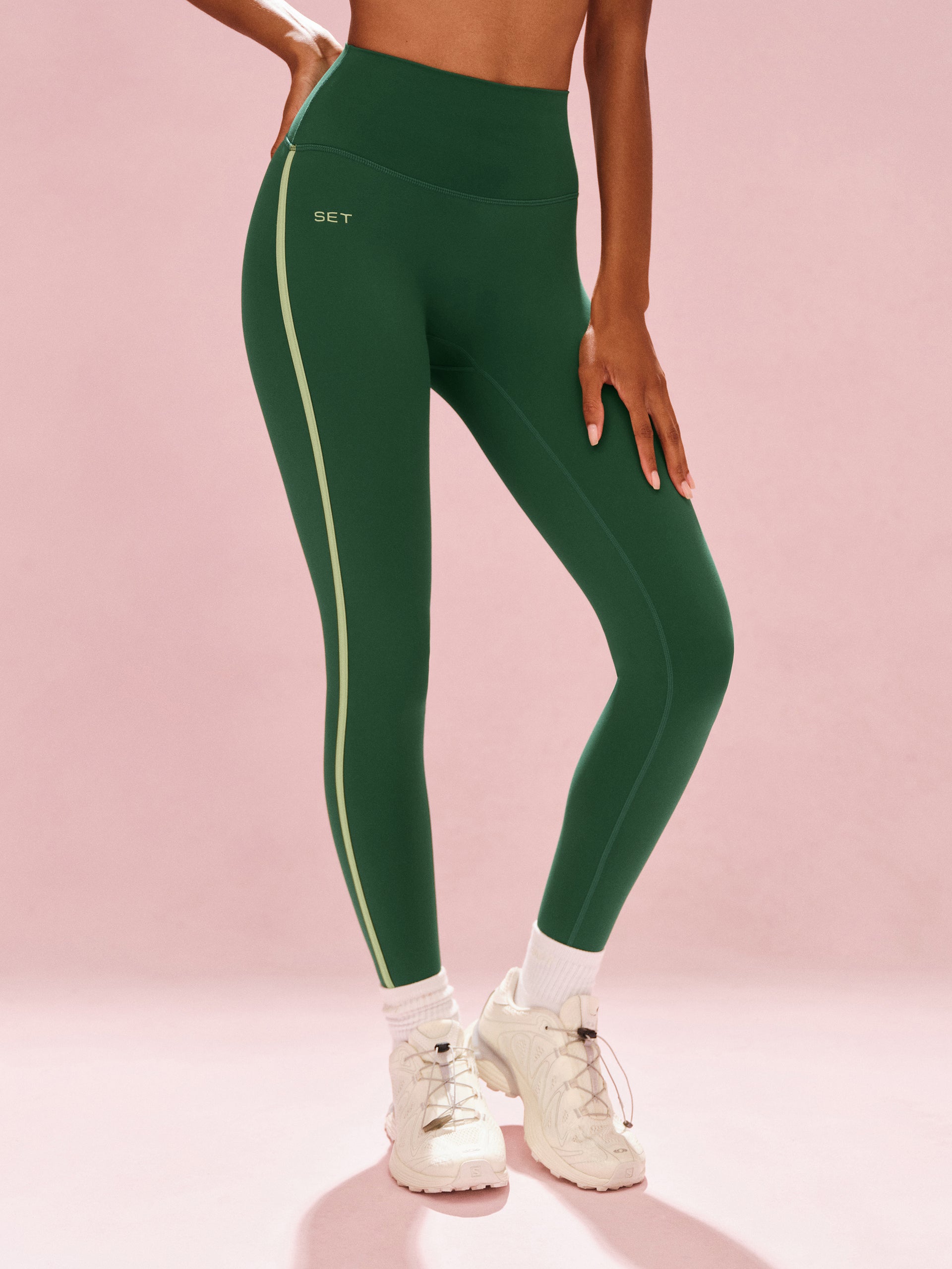 Green leggings with a brand logo on a pink background <model-info>Sydney wears a size XS and is 5’9, 24½” waist, 32B bust.</model-info>