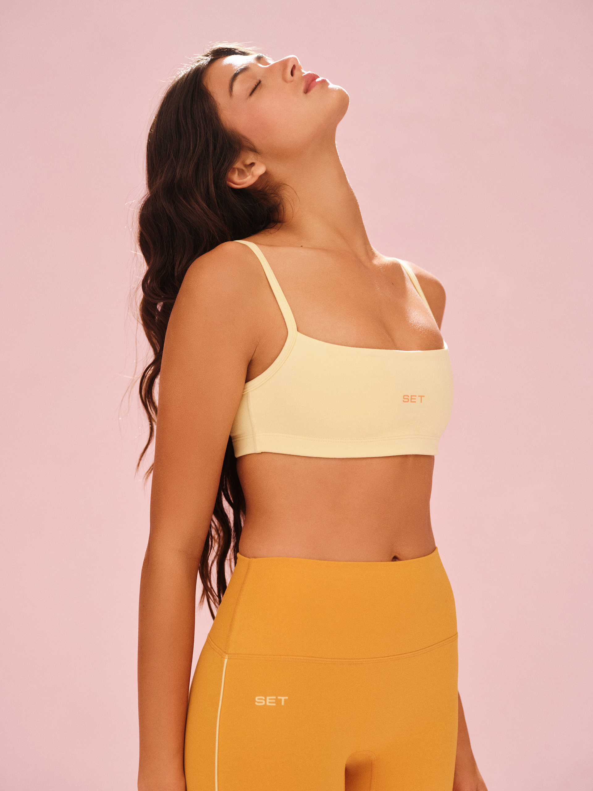 Woman wearing a yellow sports bra and mustard leggings against a pink background <model-info>Meeya wears a size XS and is 5’7, 23” waist, 32B bust.</model-info> 