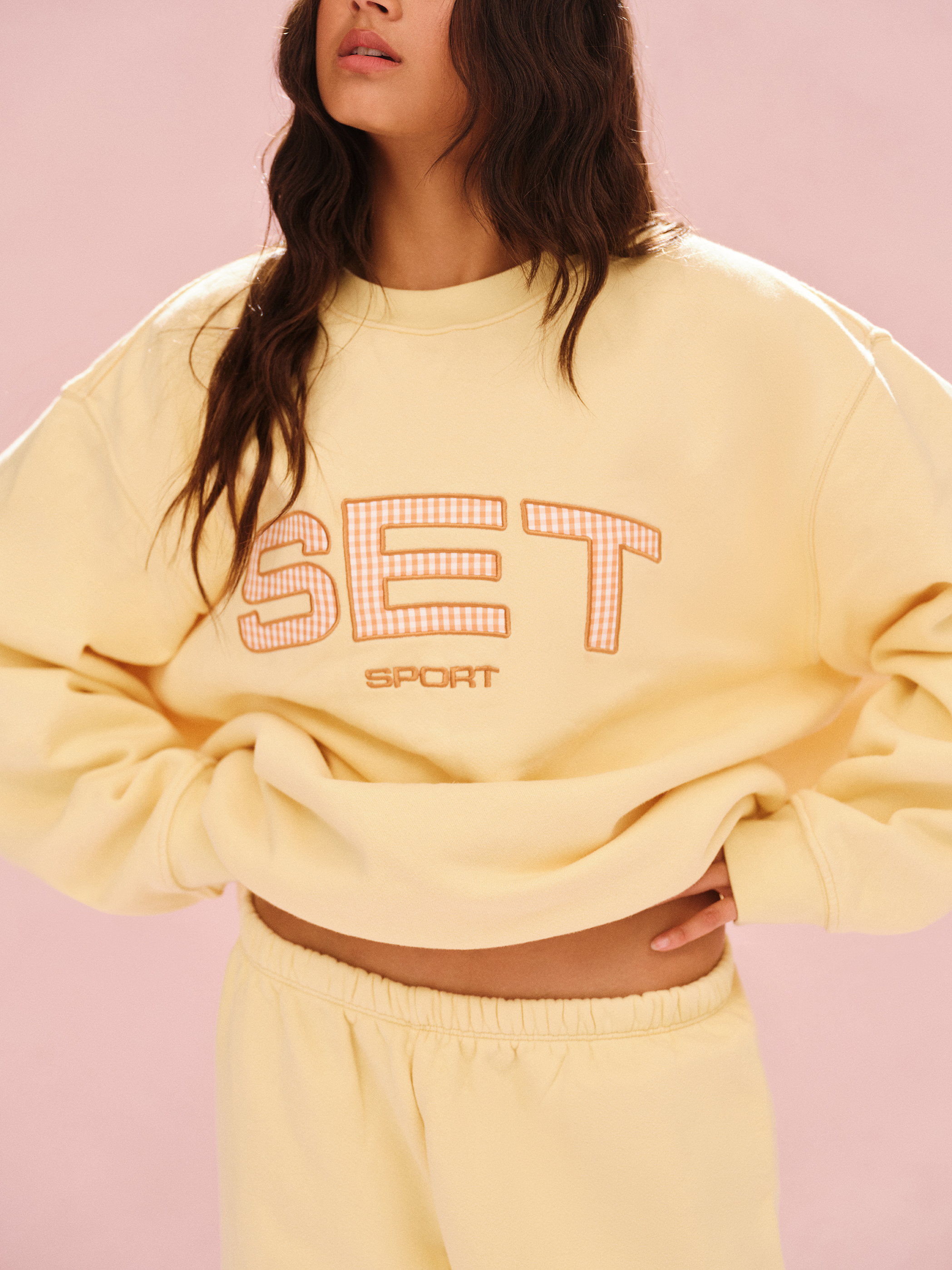 Person wearing a yellow sweatshirt with 'SET' printed on it against a pink background 