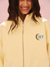 Person wearing a yellow jacket with a logo on a pink background <model-info>Meeya wears a size S and is 5’7, 23” waist, 32B bust.</model-info> 