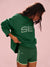 Person wearing a green 'SET' branded outfit on a pink background <model-info>Sydney wears a size M/L and is 5’9, 24½” waist, 32B bust.</model-info>