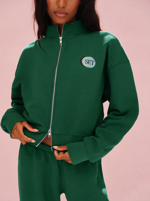 Person wearing a green tracksuit with a visible brand logo on a pink background 