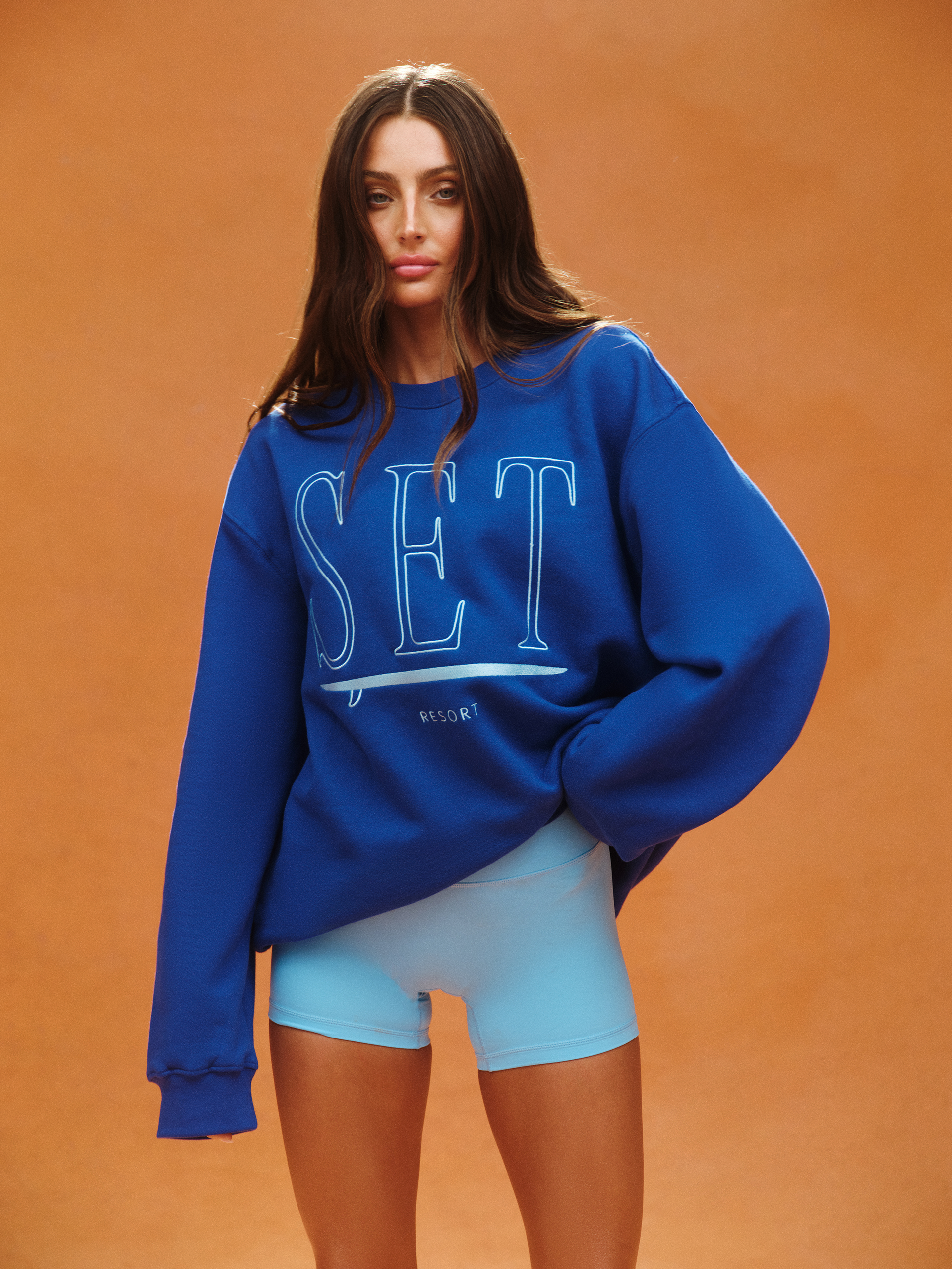 Woman wearing a blue sweatshirt with 'SET' text on an orange background <model-info>Logan wears a size M/L and is 5’9½”, 24” waist, 32B bust.</model-info>