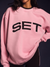 Person wearing a pink sweatshirt with 'SET' printed on it against a dark background