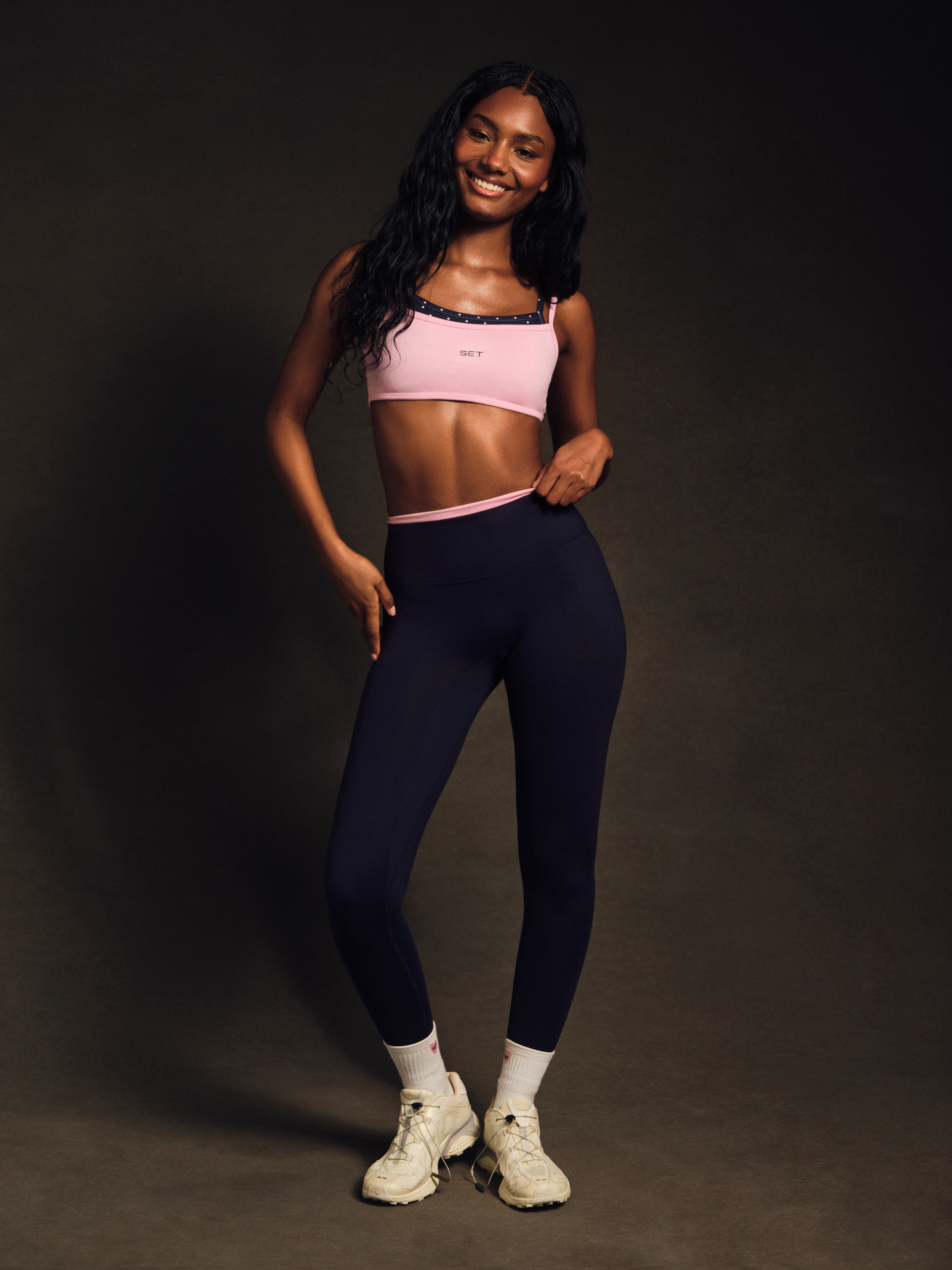 Woman wearing a pink sports bra and dark blue leggings on a dark background <model-info>Umi wears a size XS and is 5’8½”, 23½” waist, 32B bust.</model-info>