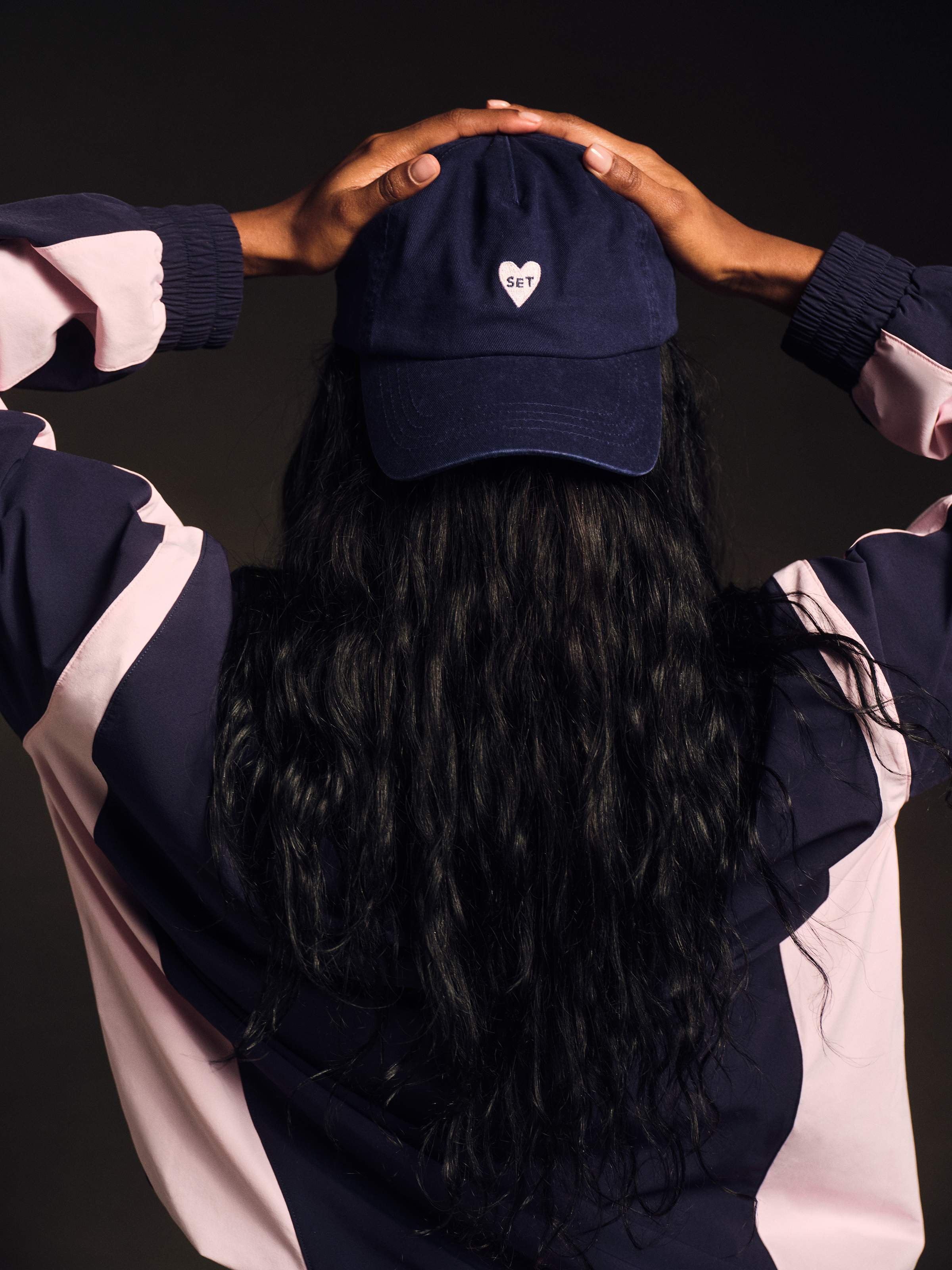Person wearing a navy blue cap with a pink heart and 'SET' logo, holding their hands on top of their head against a black background.