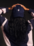 Person wearing a navy blue cap with a pink heart and 'SET' logo, holding their hands on top of their head against a black background.