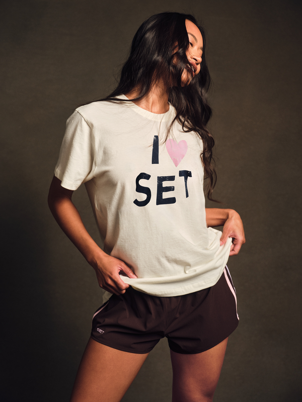 Woman wearing a white t-shirt with 'I ❤️ SET' text and dark shorts on a brown background