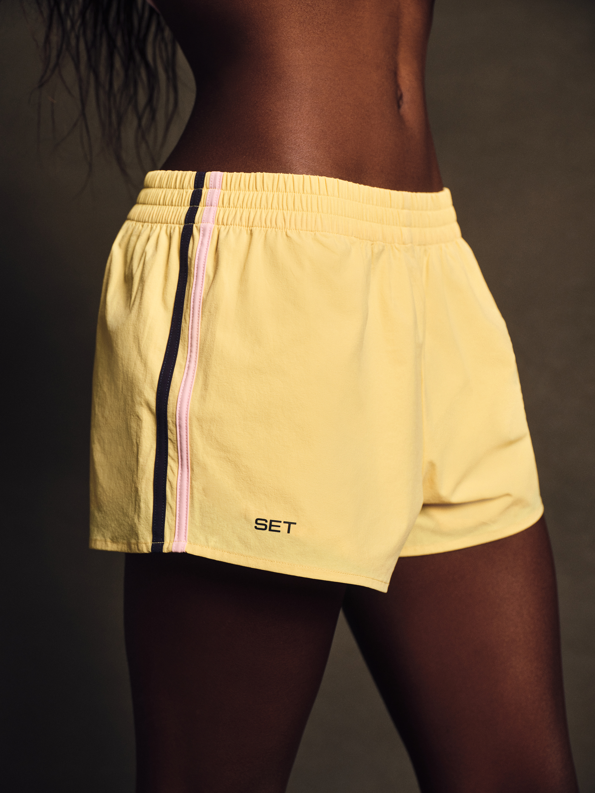 Yellow athletic shorts with a brand logo on a dark background <model-info>Nina D wears a size S and is 5’10”, 24” waist, 31½" bust.</model-info>