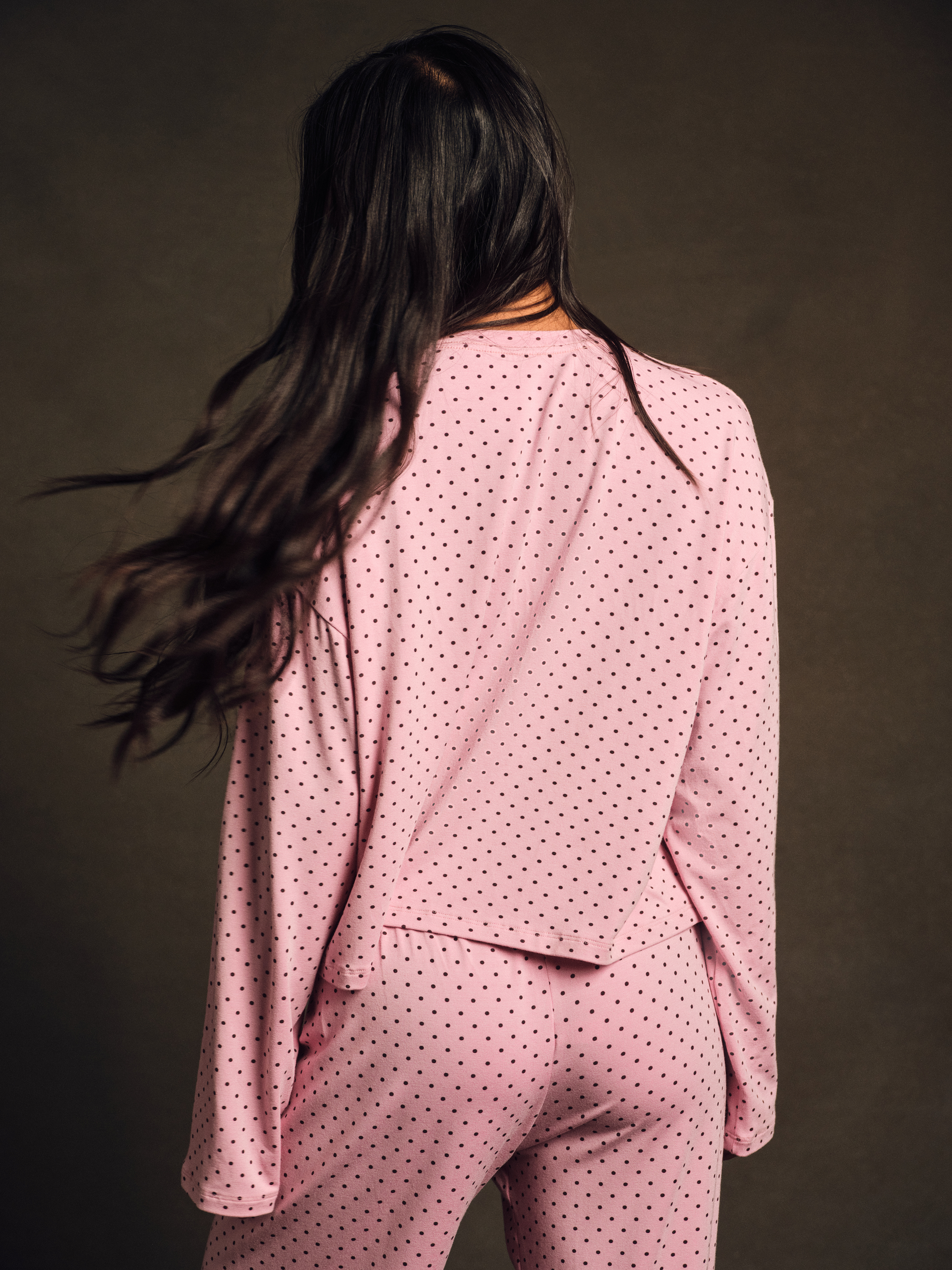 Person wearing a pink polka dot outfit with long dark hair against a brown background