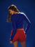 Woman wearing a blue long-sleeve top and red shorts against a dark blue background <model-info>Kushi wears a size XS and is 5'8, 24” waist, 32C bust.</model-info>