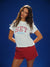 Woman wearing a white t-shirt with red and blue text against a blue background <model-info>Kushi wears a size S and is 5'8, 24” waist, 32C bust.</model-info>