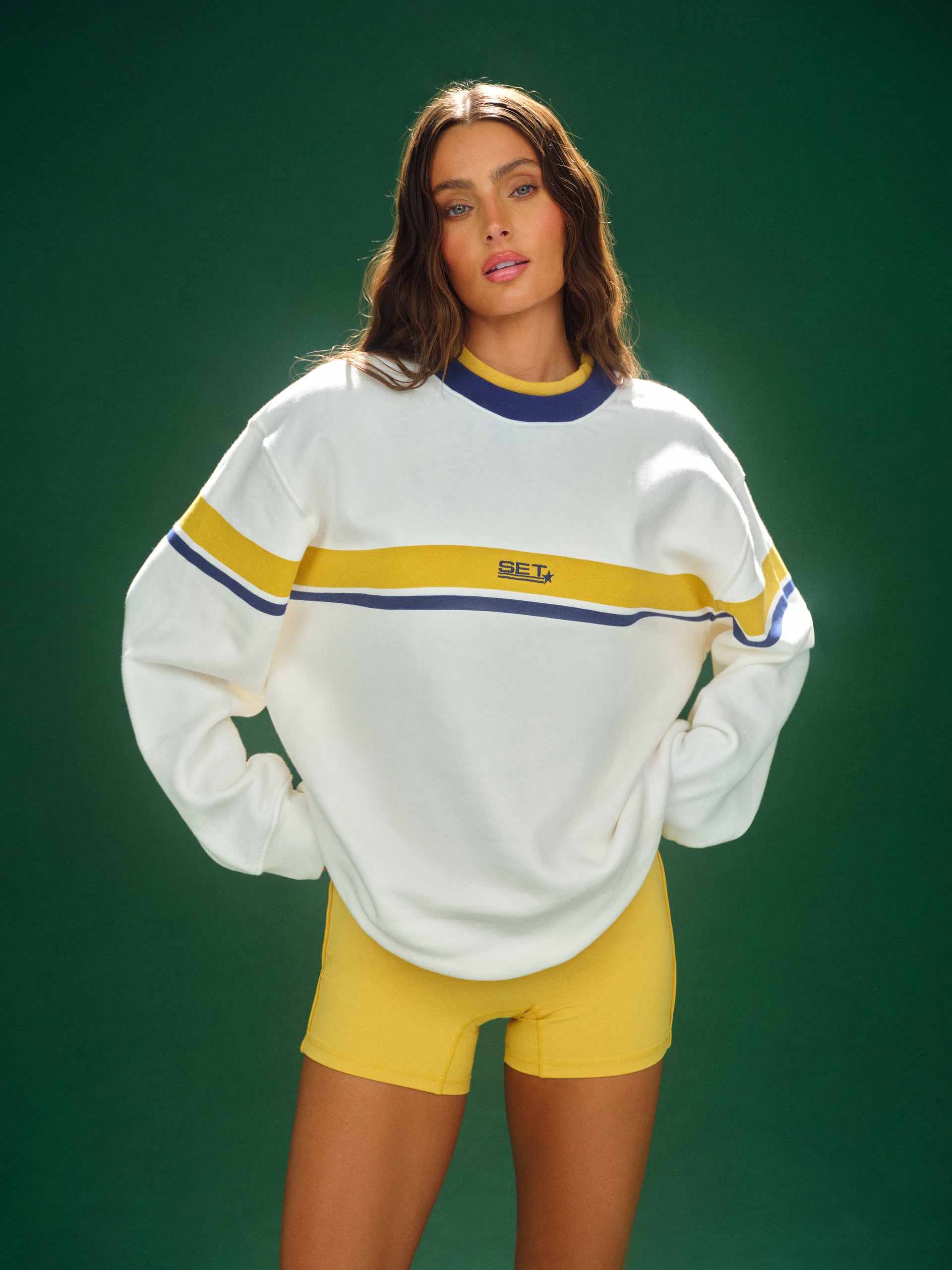 Woman wearing a white sweater with yellow and blue stripes and yellow shorts on a green background <model-info>Logan wears a size S and is 5’9½”, 24” waist, 32B bust.</model-info>