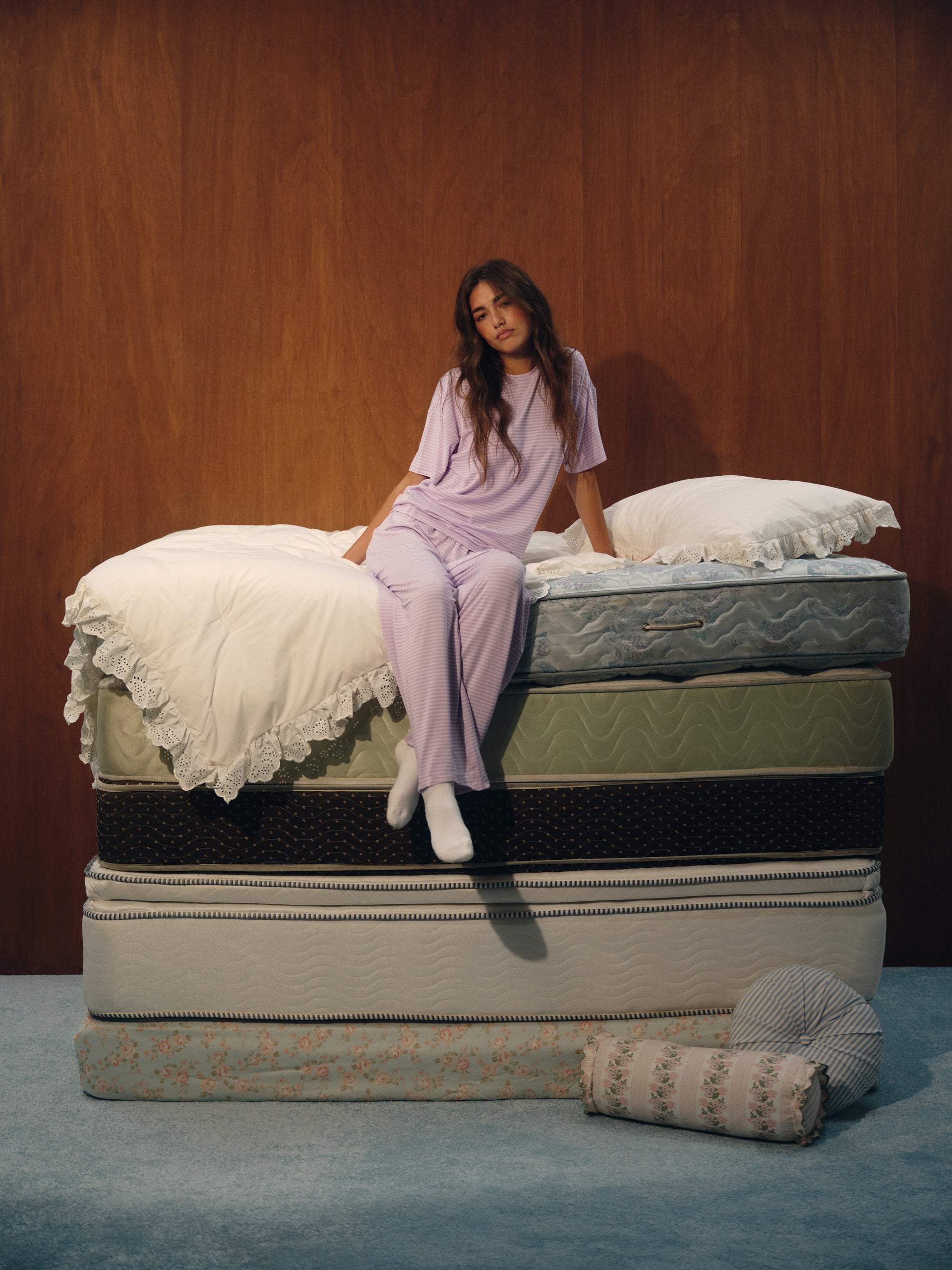 A woman in a light purple pajama set sits on top of a stacked mattress display in front of a wood-paneled wall. The mattresses are shown in various colors and textures, with white bedding on the top mattress and a rolled patterned mattress beside it. <model-info>Kalia wears a size XS and is 5’9”, 23” waist, 32B bust.</model-info>