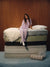 A woman in a light purple pajama set sits on top of a stacked mattress display in front of a wood-paneled wall. The mattresses are shown in various colors and textures, with white bedding on the top mattress and a rolled patterned mattress beside it. <model-info>Kalia wears a size XS and is 5’9”, 23” waist, 32B bust.</model-info>