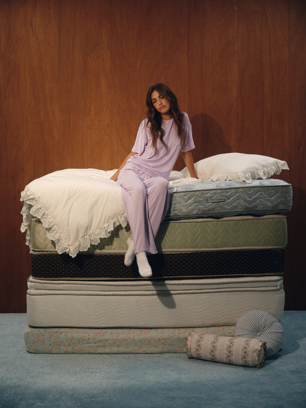 A woman in a light purple pajama set sits on top of a stacked mattress display in front of a wood-paneled wall. The mattresses are shown in various colors and textures, with white bedding on the top mattress and a rolled patterned mattress beside it. 