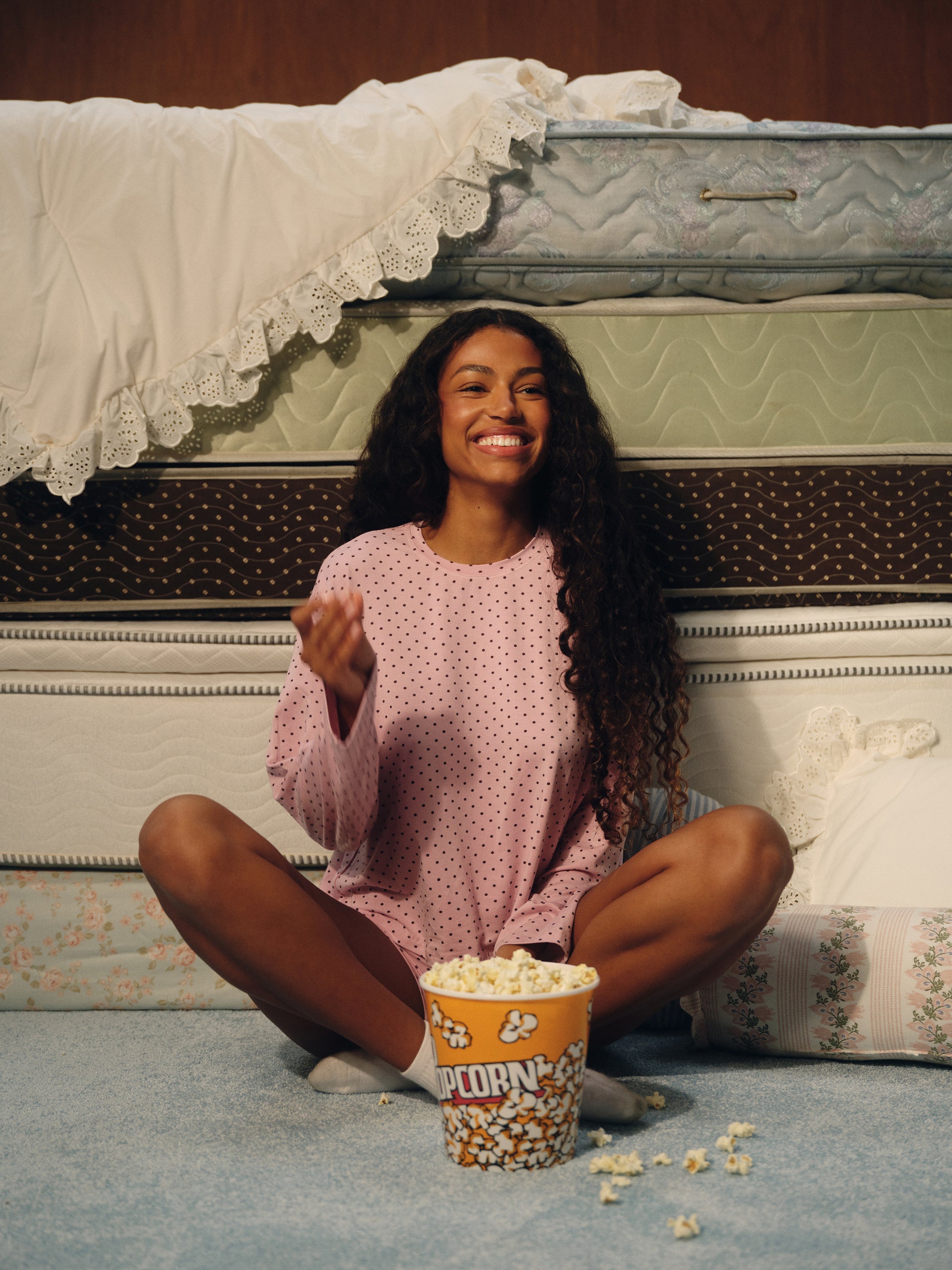 A woman with long curly dark hair sits cross-legged on a blue carpet in front of stacked mattresses, wearing a pink long-sleeve pajama top with small white polka dots. A bucket of popcorn sits in front of her.