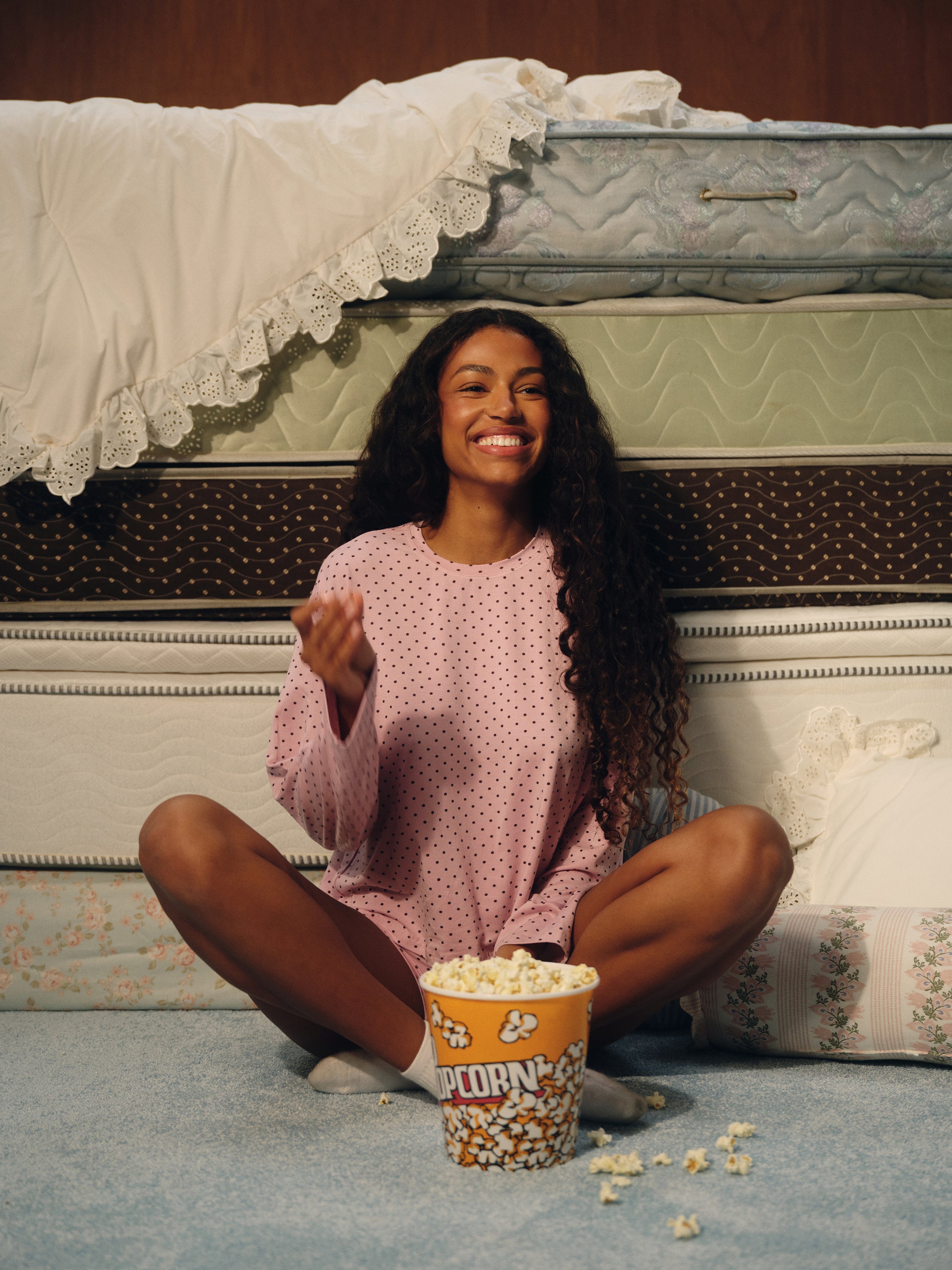 A woman with long curly dark hair sits cross-legged on a blue carpet in front of stacked mattresses, wearing a pink long-sleeve pajama top with small white polka dots. A bucket of popcorn sits in front of her.