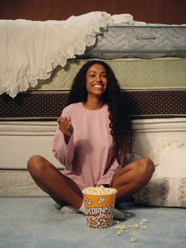 A woman with long curly dark hair sits cross-legged on a blue carpet in front of stacked mattresses, wearing a pink long-sleeve pajama top with small white polka dots. A bucket of popcorn sits in front of her.