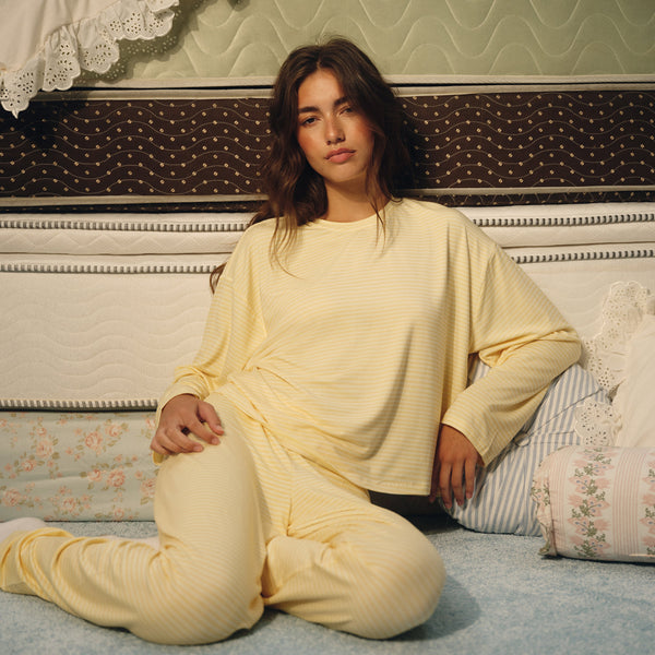 A woman sits on a light blue carpet in front of a bed, wearing a pale yellow ribbed lounge set consisting of a long-sleeve top and matching pants.