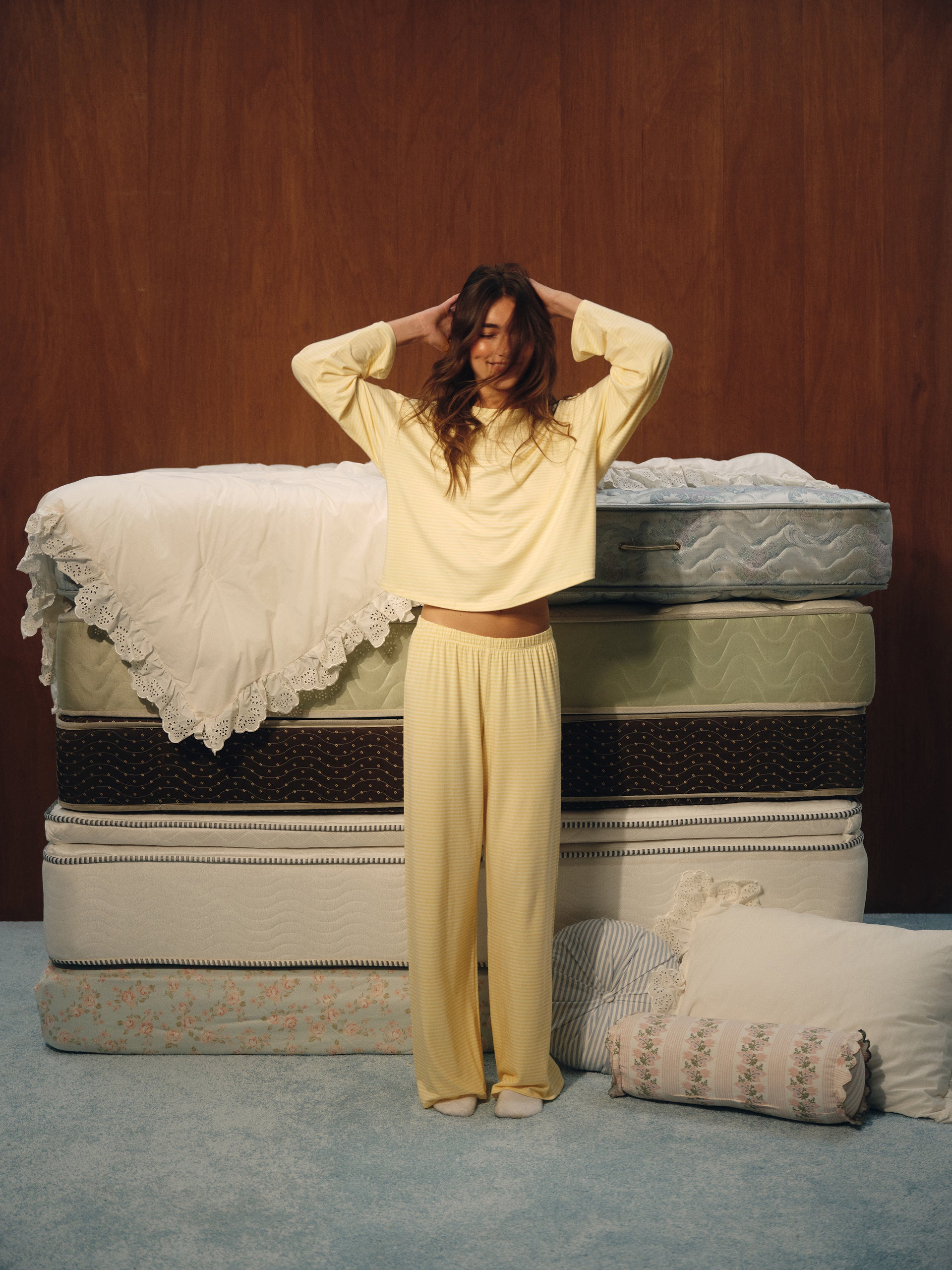 A woman wearing a pale yellow long-sleeve cropped top and matching wide-leg pajama pants stands in front of a stack of mattresses with pillows on a blue carpeted floor.