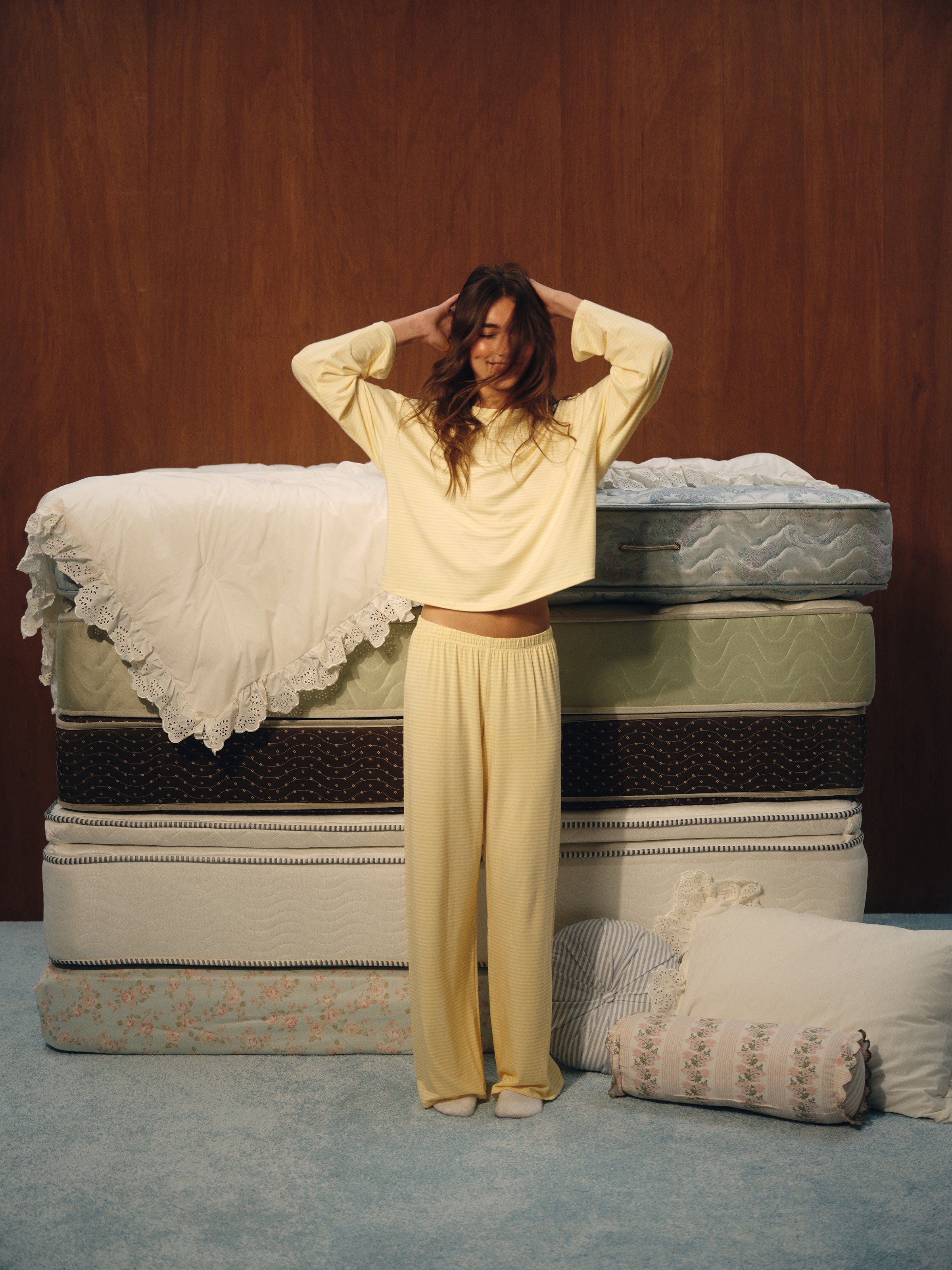 A woman wearing a pale yellow long-sleeve cropped top and matching wide-leg pajama pants stands in front of a stack of mattresses with pillows on a blue carpeted floor.