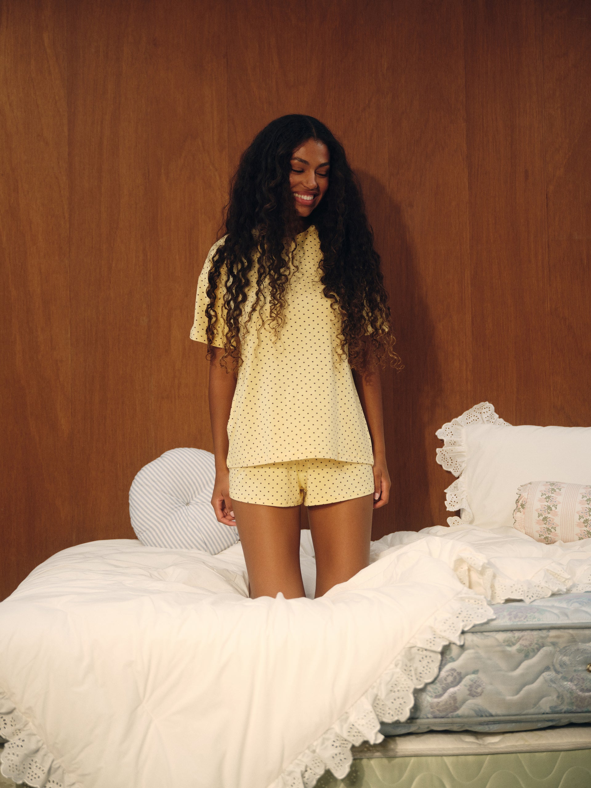 A woman with long curly black hair stands on a bed wearing a pale yellow short-sleeve pajama set with small white polka dots. The bedroom has a warm wood-paneled wall, white bedding, and a light blue quilted mattress.
