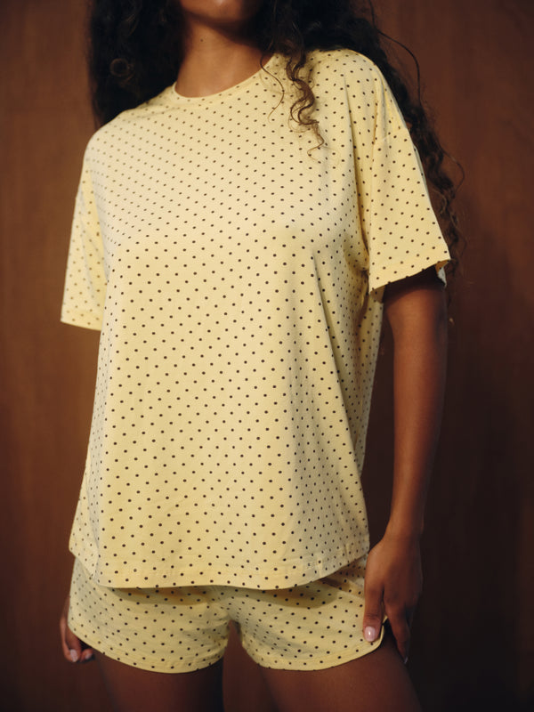 A woman wearing a pale yellow short-sleeve t-shirt and matching shorts set with a small black polka dot pattern, set against a dark brown wooden background.
