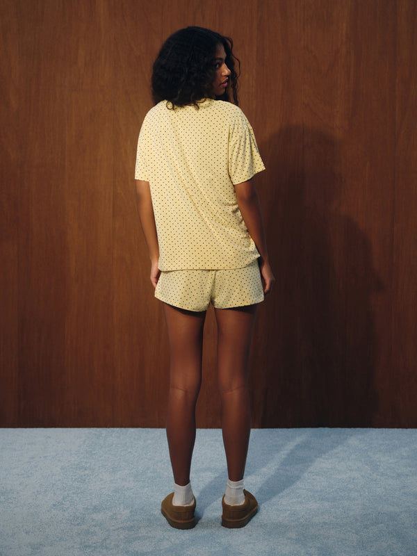 A woman stands with her back to the camera wearing a light yellow short-sleeve t-shirt and matching shorts with a small white polka dot pattern, paired with white socks and brown shoes, set against a dark wood-paneled wall and a light blue carpet.
