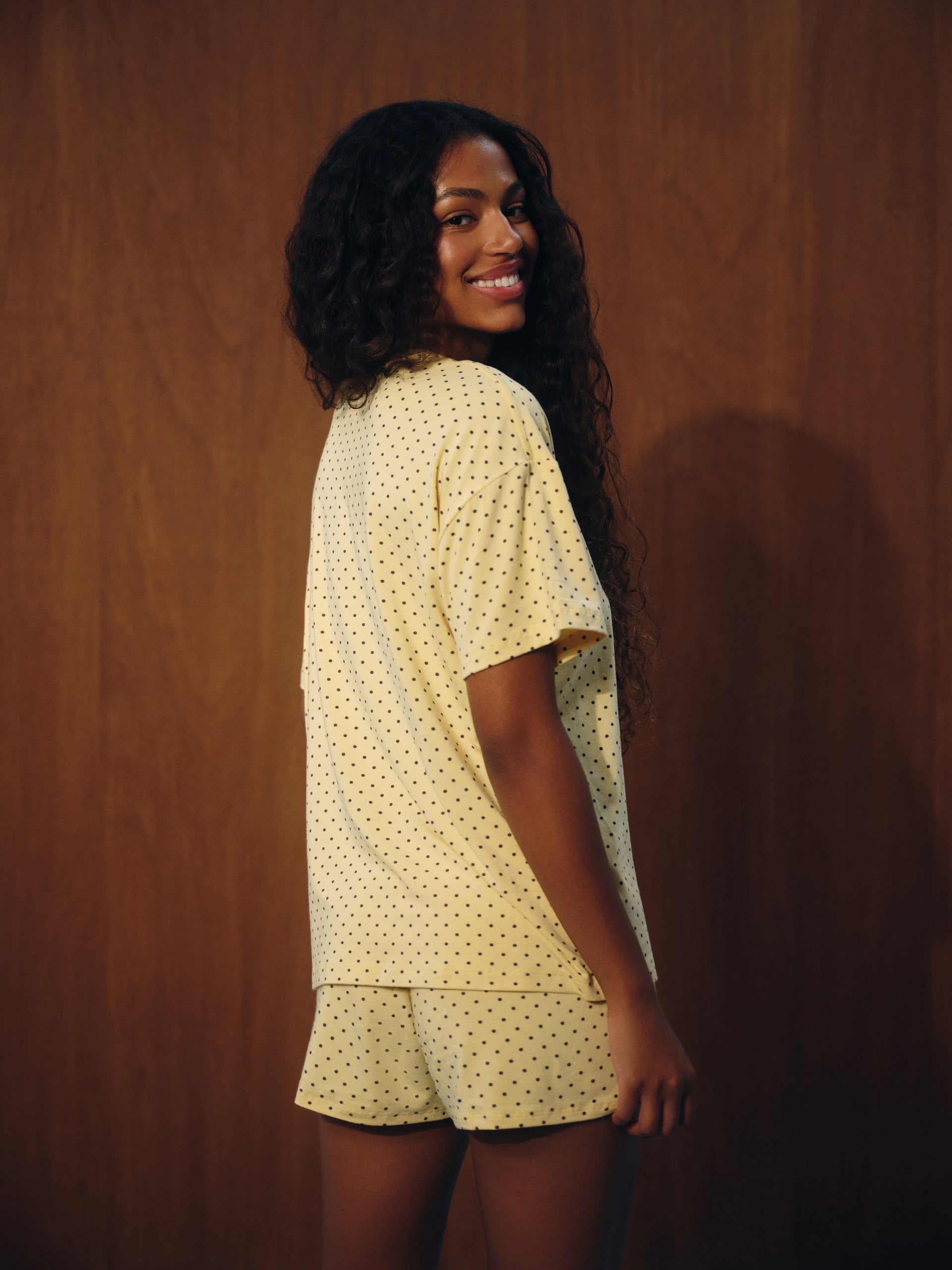 A woman with long curly black hair is shown from the back wearing a pale yellow short-sleeve pajama top and matching shorts with a small black polka dot pattern, set against a dark wood-paneled background.
